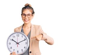 Beautiful caucasian woman with blonde hair wearing business jacket and holding clock smiling happy pointing with hand and finger Beautiful caucasian woman with blonde hair wearing business jacket and holding clock smiling happy pointing with hand and finger
