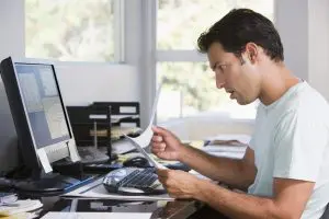 Man in home office using computer holding paperwork and looking